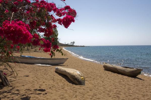 A thatched gazebo on a stone pier over Lake Malawi's turquoise water, with a traditional sailboat passing a rocky island